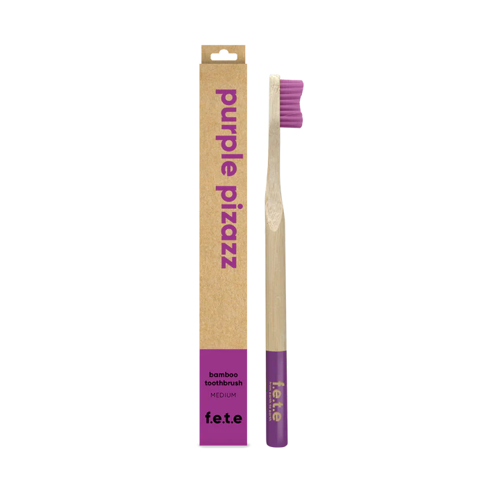 A F.E.T.E. Single Medium Bamboo Toothbrush with a biodegradable handle and purple bristles is shown next to its "purple pizazz" box, available at Fisa Health.