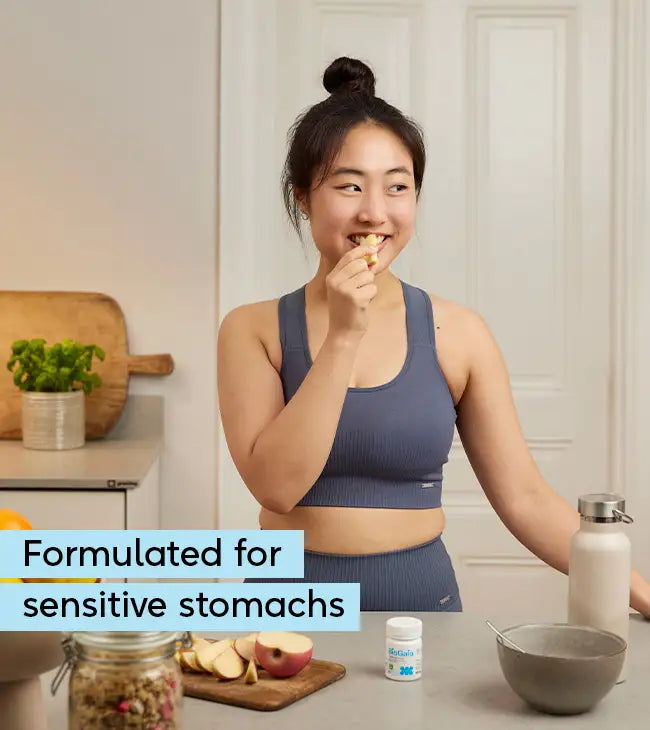 A woman in workout clothes stands in her kitchen eating an apple next to BioGaia® Gastrus® PURE, with fruit and a glass of water on the counter, packaging displayed, available at Fisa Health.