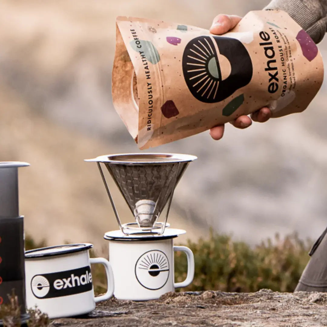 A hand pours Exhale Coffee - House - Ground (Medium) into a metal pour-over on a white mug, with another Fisa Health branded mug beside it outdoors, available at Fisa Health.