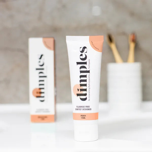 A tube of Dimples Hydroxyapatite Toothpaste (Peach) is displayed on a white surface, with its box and two toothbrushes softly out of focus in the background, available at Fisa Health.