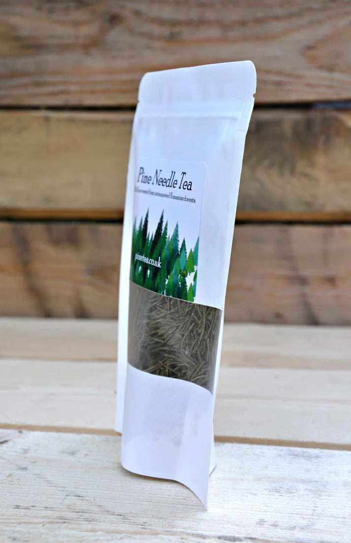 A white, resealable bag labeled Pinetea Pine Needle Tea with a green forest design is displayed on a light wooden surface against a wooden background, available at Fisa Health.