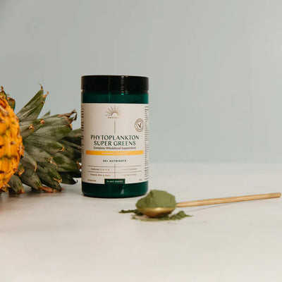 A jar of Phytoplankton Super Greens Powder 90g rests on a white surface beside a wooden spoon filled with green powder and a pineapple slice, available at Fisa Health.