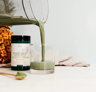 A blender pours a green smoothie into a glass next to Phytoplankton Super Greens Powder 90g, a spoon with powder, and a pineapple on a white surface, available at Fisa Health.