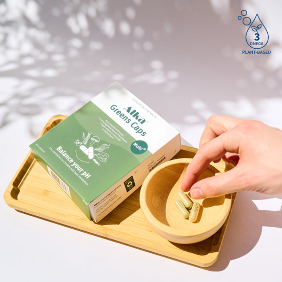 A hand picks a green capsule from a bowl next to Alka® Greens Multi - Capsules, with icons representing nutrients, immune support, and plant-based omega-3s, available at Fisa Health.