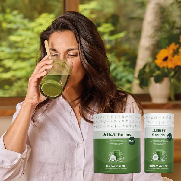 A woman drinks a green smoothie in a sunlit kitchen, with two containers of Alka® Greens Multi superfood powder visible on the counter, available at Fisa Health.