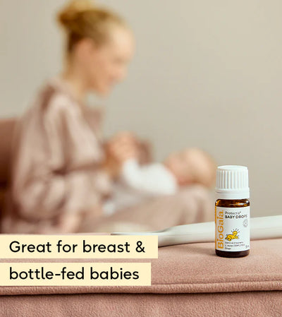A bottle labeled "BioGaia Protectis Baby Drops 5ml" by is on a surface, with a blurred woman in a robe holding her baby in the background. Available at Fisa Health.