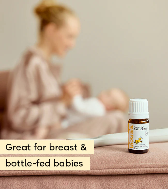 A bottle labeled "BioGaia Protectis Baby Drops 5ml" by is on a surface, with a blurred woman in a robe holding her baby in the background. Available at Fisa Health.