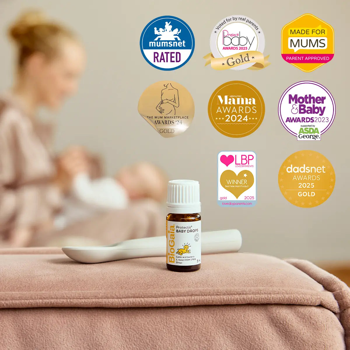 A bottle labeled BioGaia Protectis Baby Drops 5ml is placed on a soft blanket next to a spoon, with award badges above. A mother is seen feeding her baby. Available at Fisa Health.