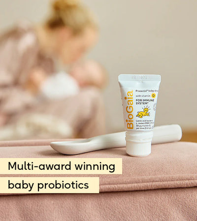 A bottle of BioGaia Protectis Baby Drops with Vitamin D 10ml and a white spoon on a pink surface, with a blurred mother and baby in the background, available at Fisa Health.