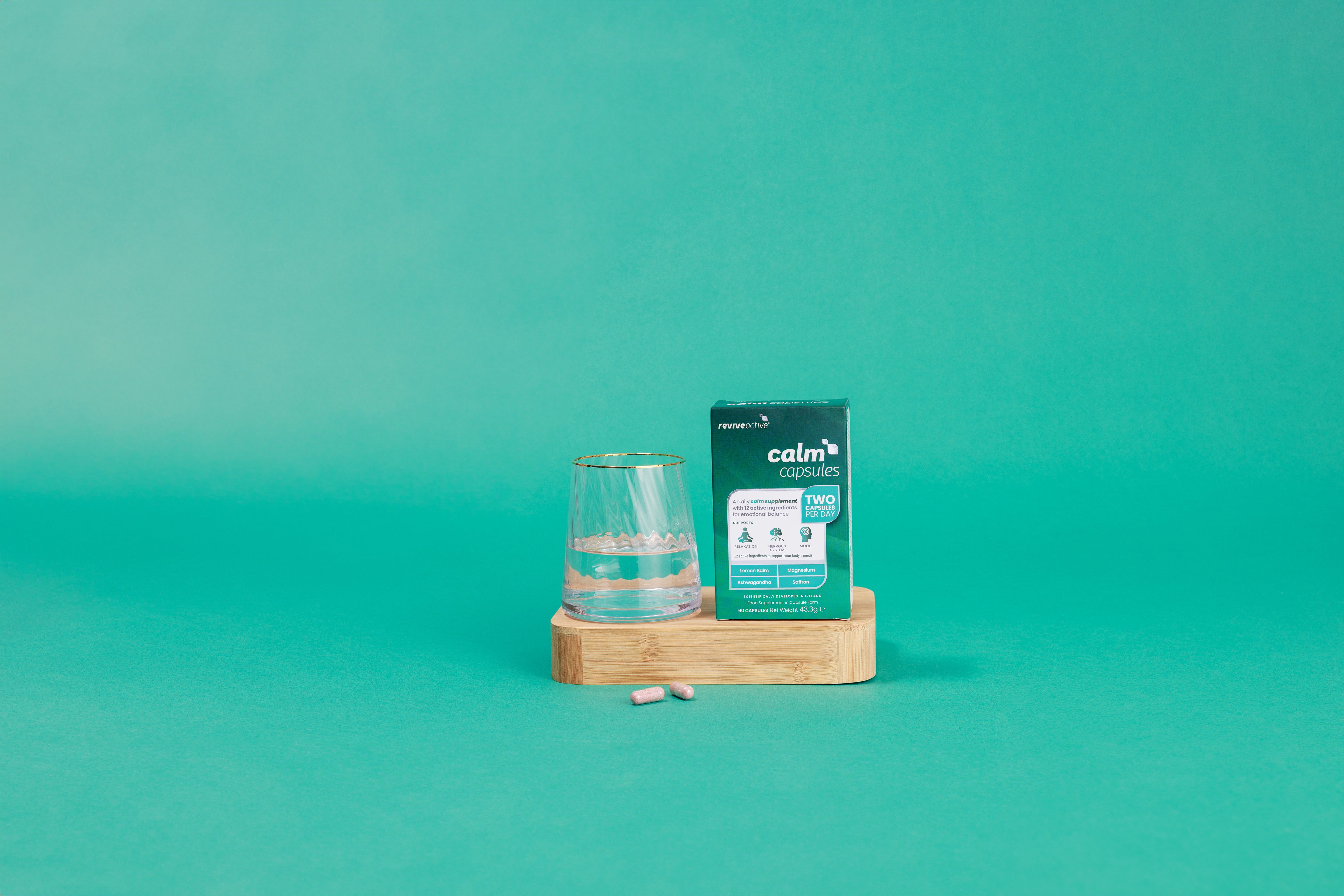 A glass of water and a container labeled "Revive Active Calm Capsules" are on a wooden tray with two pink capsules, set against a turquoise background, available at Fisa Health.