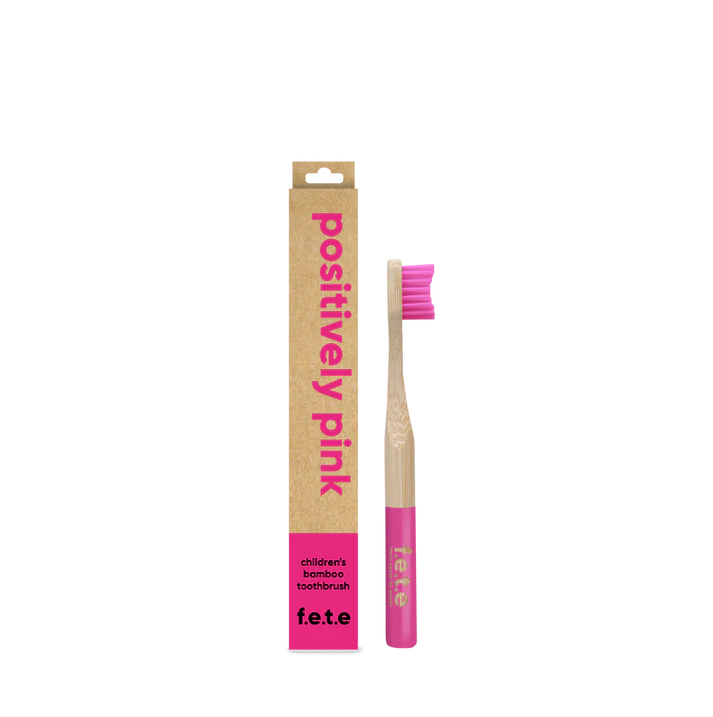 A pink and natural bamboo F.E.T.E. Children’s Bamboo Toothbrush stands upright beside its “positively pink” labeled cardboard box, available at Fisa Health.