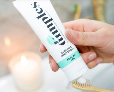 A hand squeezes Dimples Hydroxyapatite Toothpaste onto a wooden toothbrush, with a blurred lit candle in the background, available at Fisa Health.