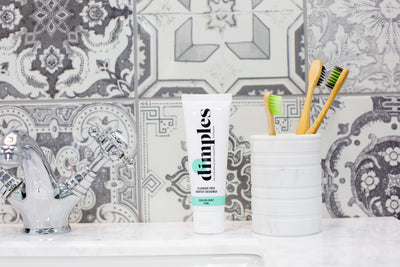 A bathroom sink with Dimples Hydroxyapatite Toothpaste, a marble cup holding three bamboo toothbrushes, and a tube of shave cream on patterned tile, available at Fisa Health.