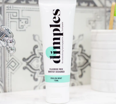 A tube of Dimples Hydroxyapatite Toothpaste sits on a bathroom counter, with patterned tiles in the background, available at Fisa Health.