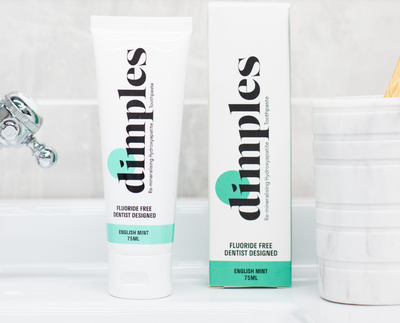 A tube and box of Dimples Hydroxyapatite Toothpaste English Mint are placed on a bathroom sink beside a marble toothbrush holder, available at Fisa Health.