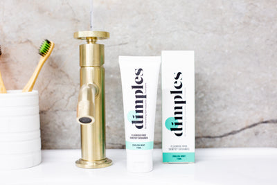 A tube and box of Dimples Hydroxyapatite Toothpaste are placed next to a gold faucet and toothbrush holder on a bathroom sink with a stone wall background, available at Fisa Health.