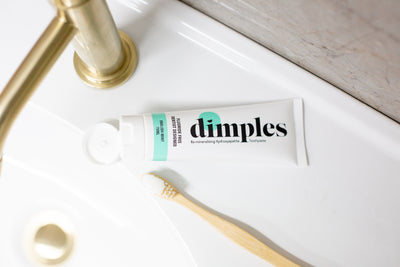 A tube of Dimples Hydroxyapatite Toothpaste with an open cap is on a white sink next to a bamboo toothbrush, near a brass faucet, available at Fisa Health.