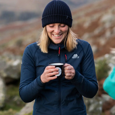 A woman in a navy jacket and beanie smiles outdoors, holding a mug of Exhale Coffee - House - Ground (Medium); blurred hills in background. Available at Fisa Health.