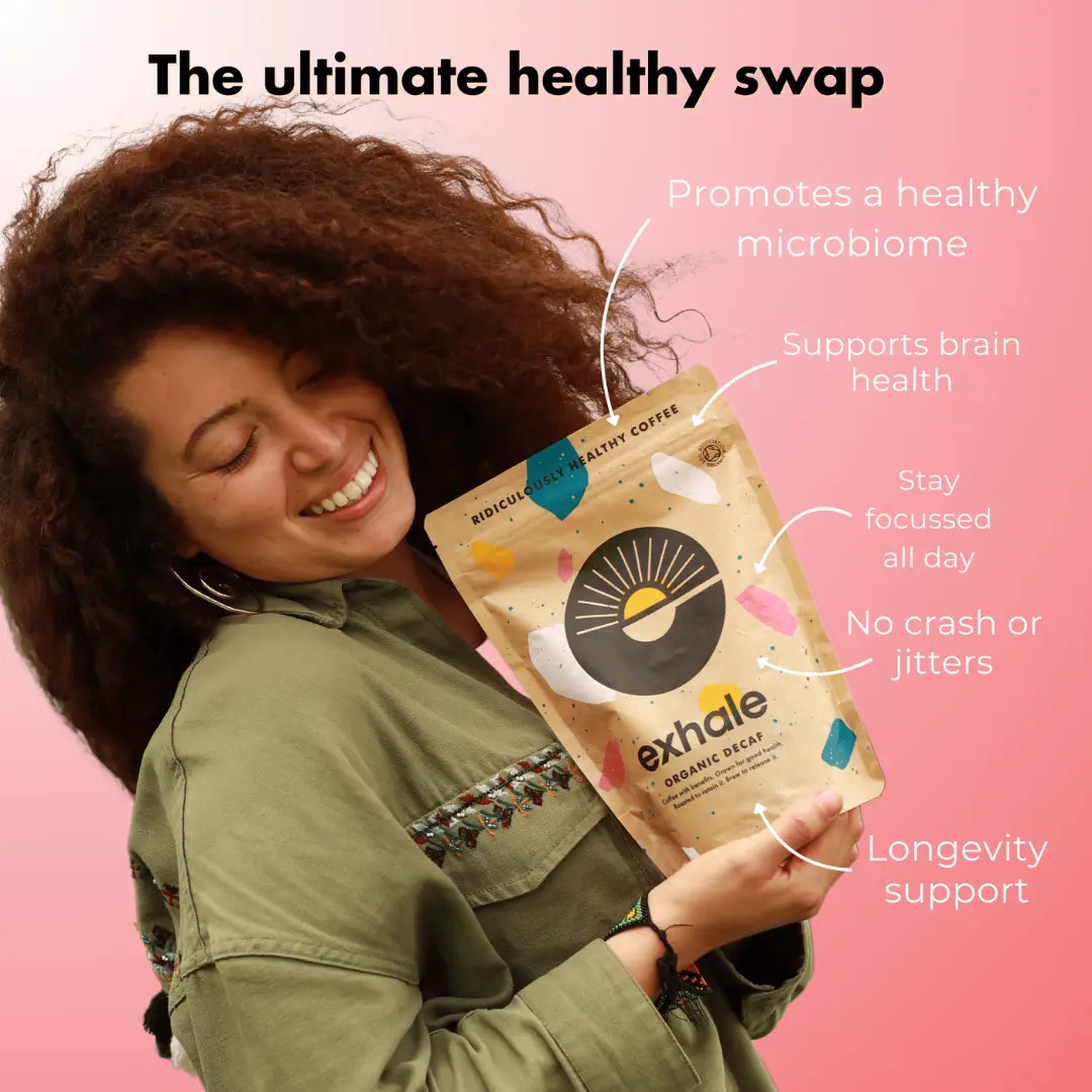 A smiling woman in a green shirt hugs a bag of Exhale Coffee Decaf - Ground (Medium) with the product name visible on the packaging, set against a pink background; available at Fisa Health.