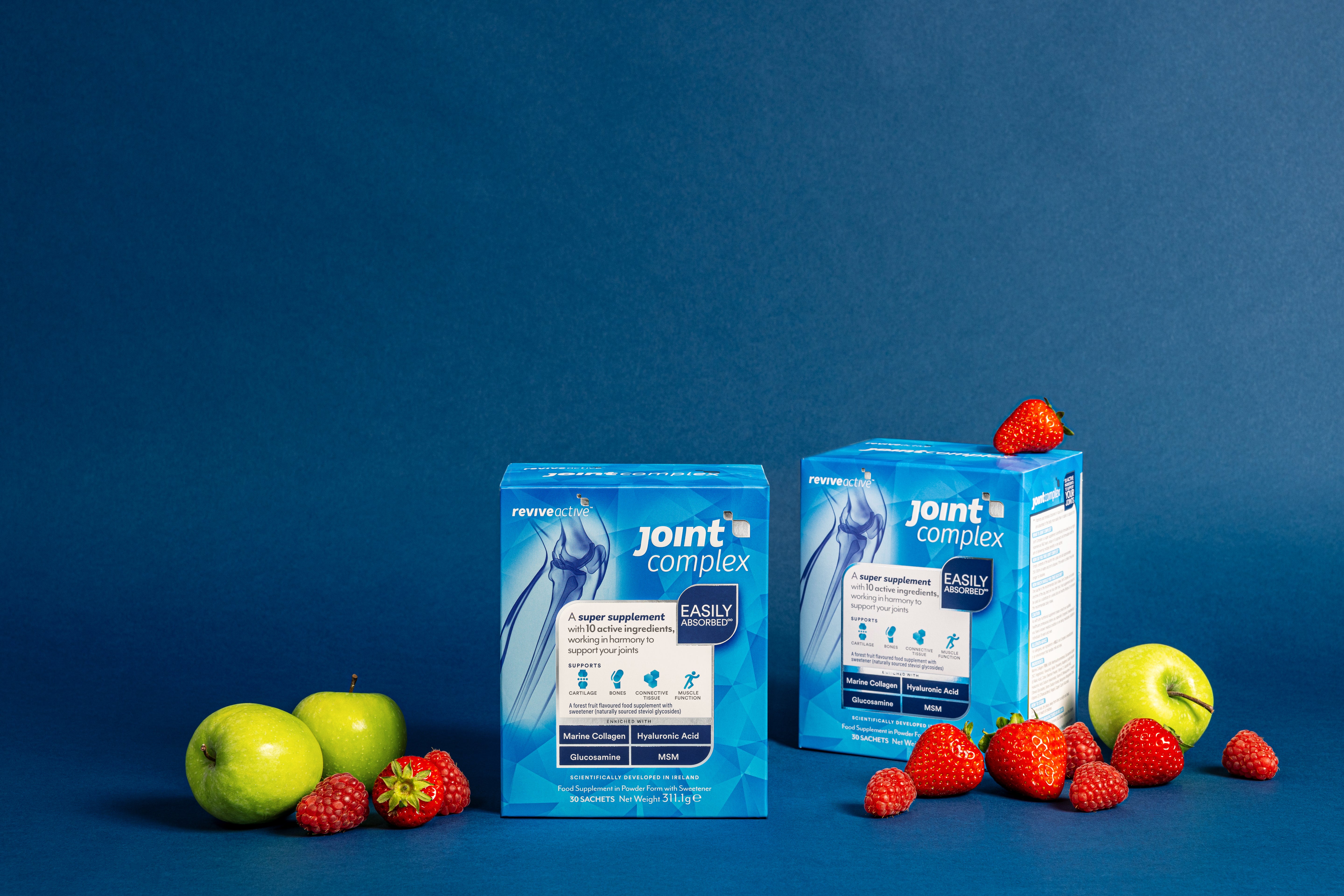 Two blue boxes of Revive Active Joint Complex are displayed with green apples, strawberries, and raspberries on a blue background, available at Fisa Health.
