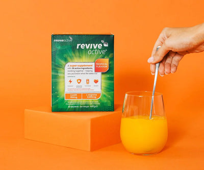 A hand stirs an orange beverage in a glass with a metal spoon next to a box of Revive Active O+M Orange & Mango Multi-Nutrient Powder, set on an orange background, available at Fisa Health.
