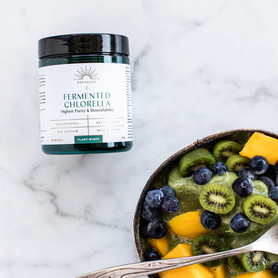 A jar labeled "Fermented Chlorella Powder" is on a marble surface next to a smoothie bowl with blueberries, kiwi, and mango. Available at Fisa Health.