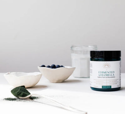 A jar labeled "Fermented Chlorella Powder" is on a white table with a spoonful of powder, blueberries, and a glass of water in the background; available at Fisa Health.
