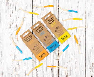 Three F.E.T.E. Bamboo Interdental Brushes and several unboxed yellow and blue handled brushes displayed on a white wooden surface, available at Fisa Health.