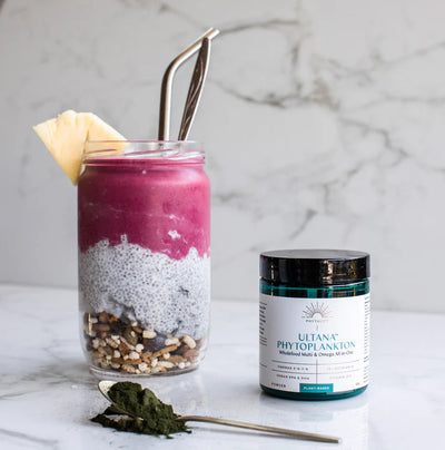 A jar with layers of granola, chia pudding, and pink smoothie topped with pineapple sits next to Phytoplankton Powder 60g Omega & Multi and a spoonful of powder, available at Fisa Health.