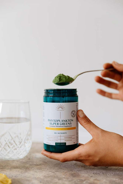 A hand holds a jar of Phytoplankton Super Greens Powder 90g while another uses a scoop by a glass of water on the table, available at Fisa Health.