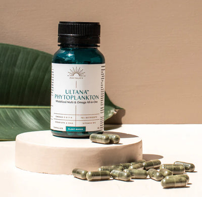 A bottle of Phytoplankton 60 Veg. Capsules (500mg) Omega & Multi sits on a beige platform with green capsules and a large leaf in the background, available at Fisa Health.