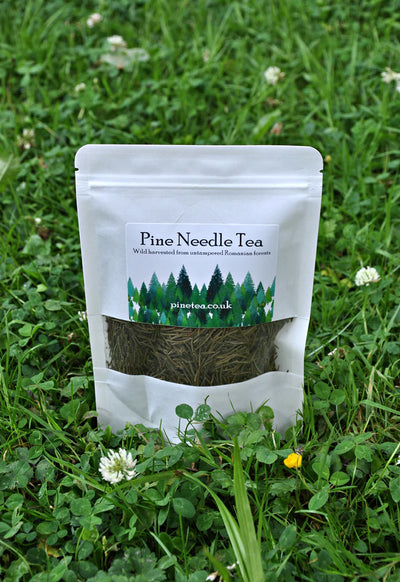 A white pouch labeled Pinetea rests on green grass with clover and small white flowers; a clear window in the packaging shows dried pine needles inside, available at Fisa Health.