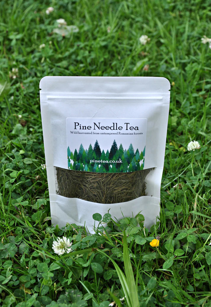 A white pouch labeled Pinetea rests on green grass with clover and small white flowers; a clear window in the packaging shows dried pine needles inside, available at Fisa Health.