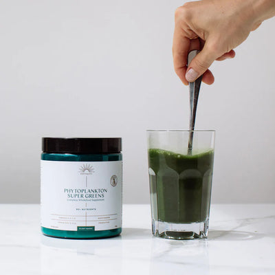 A hand stirs a glass of green drink next to a Phytoplankton Super Greens Powder 180g container on a white surface, available at Fisa Health.