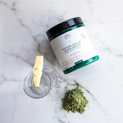 A jar labeled "Fisa Health Phytoplankton Super Greens Powder 180g," green powder, a clear glass, and a pineapple wedge on a marble surface; available at Fisa Health.