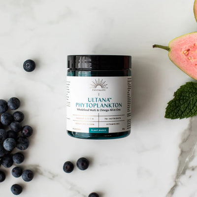 A jar of Phytoplankton Powder 60g Omega & Multi sits on a marble surface with blueberries and sliced pink guava, available at Fisa Health.